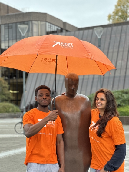 Two UL First Seven Weeks student guides in orange tshirts holding an orange umbrella over the statue of Brown Thomas 