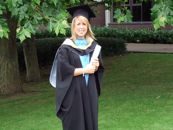 A female college graduate in black cap and robe standing on grass in front of trees holding their diploma
