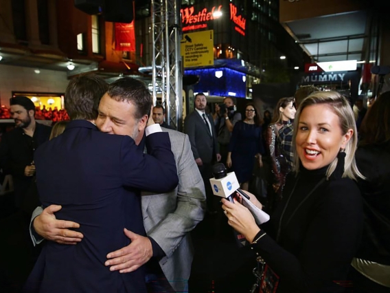 Actors Tom Cruise and Russell Crowe hugging on the red carpet as a blonde reporter holds a microphone beside them
