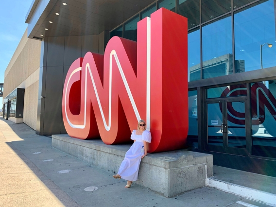 A blonde woman wearing sunglasses and a white dress sitting in front of a building with a large CNN sign