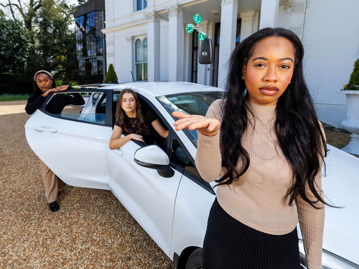 A woman standing in front of a white car throwing three green dice in the air while two women stand in the background, one sitting in the car and the other leaning on the back door
