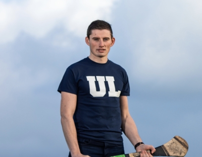 Gearoid Hegarty in a navy t-shirt with UL in white on the front.  He is holding a hurley in both hands and looking directly at the camera with clear sky behind him.