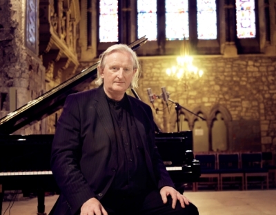 Prof Mícheál Ó Súilleabháin sits by a piano in a church setting 