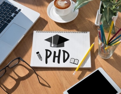stock image of a laptop and notepad with PHD written on it and a graduation cap on a desk.