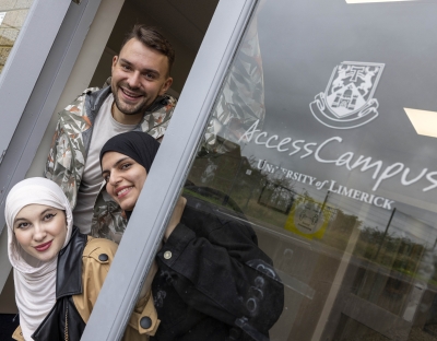 Two smiling female students wearing hijabs and a male student peaking out from the glass door with the sign UL Access Campus 