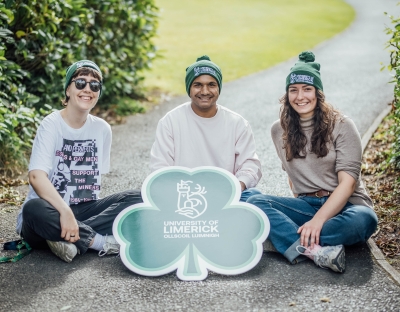 3 students wearing UL green beanies sitting on the ground with a shamrock sign with the UL logo on it