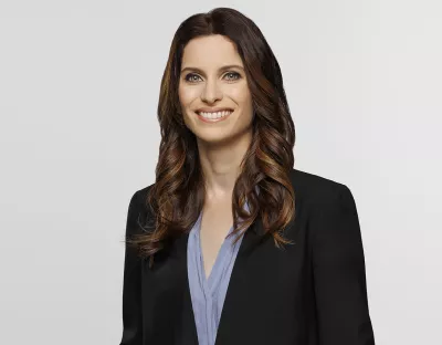 A corporate headshot of a woman with wavy brown hair wearing a black suit jacket