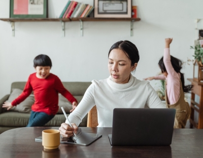 Image depicting a woman at a home work desk with two children playing in the background. 