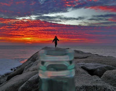 Picture of a little girl running to the end of a rocky jetty by the sea. A glass bottle appears at the base of the image making it look like she is coming out of the mouth of the bottle. A red, orange and purple sun sets through the clouds into the surrounding sea in the background.
