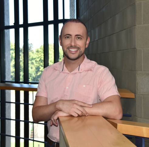 Dr Daniel Granato leaning on a wooden banister, wearing a salmon coloured shirt and smiling into the camera