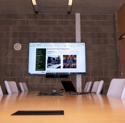 A photo of a hybrid meeting room. The photos shows a large table surrounded by chairs with a laptop resting on the table. On the wall behind the table in a large projector screen which is attached to the laptop and sharing what's on the laptop screen. 