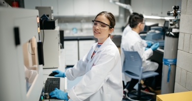A women works in a lab environment. She has a lap coat and glasses. In the background two other people are also working in the lap at a different machine.