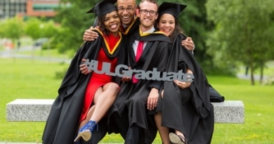 Group of four people in graduation robes and caps sitting on bench on campus holding the words hashtag U.L graduate
