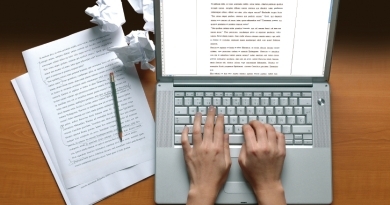 Hands typing on a laptop displaying text with sheets of text next to it, as well as crumpled up paper and a pen
