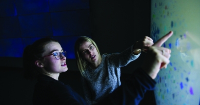 Two women looking and pointing at a large screen with data points logged in the white projector