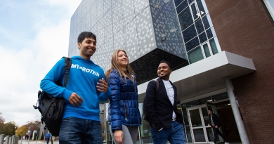 Two men and a woman walking in front of a tall building with glass panels. The trio are smiling and chatting