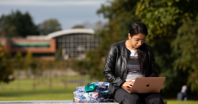 Girl sitting on a stone bench outside working on her laptop. In the background there are a row of trees in front of a large building