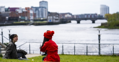 Two students sit on a green wearing winter clothes with Limerick City and the River Shannon in the background