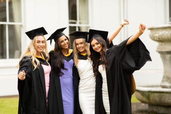 four female graduates of the UL Masters Apprenticeship Programme standing together wearing caps and gowns. In the background there is a white house.