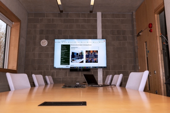 A photo of a hybrid meeting room. The photos shows a large table surrounded by chairs with a laptop resting on the table. On the wall behind the table in a large projector screen which is attached to the laptop and sharing what's on the laptop screen. 