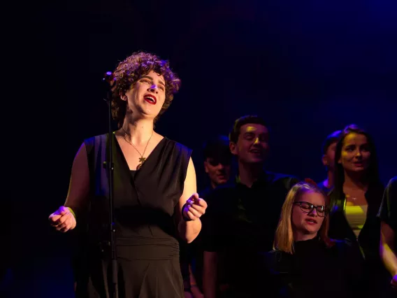 A person in a black top singing on stage with other performers in the background