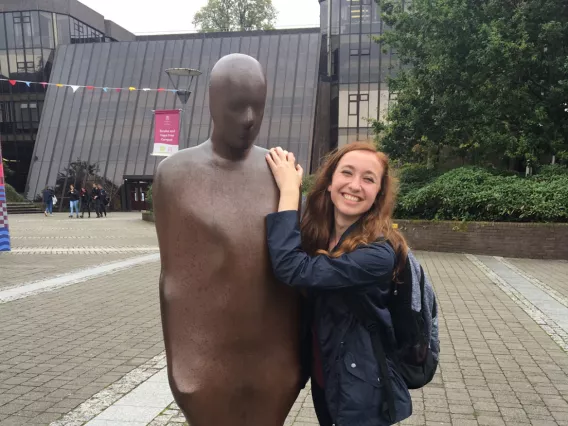 A woman with brown hair and a navy jacket posing with a sculpture of a man