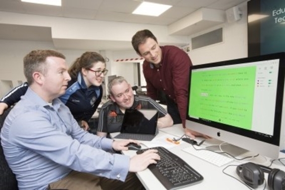 A man sitting at a computer demonstrating software to three people looking on.