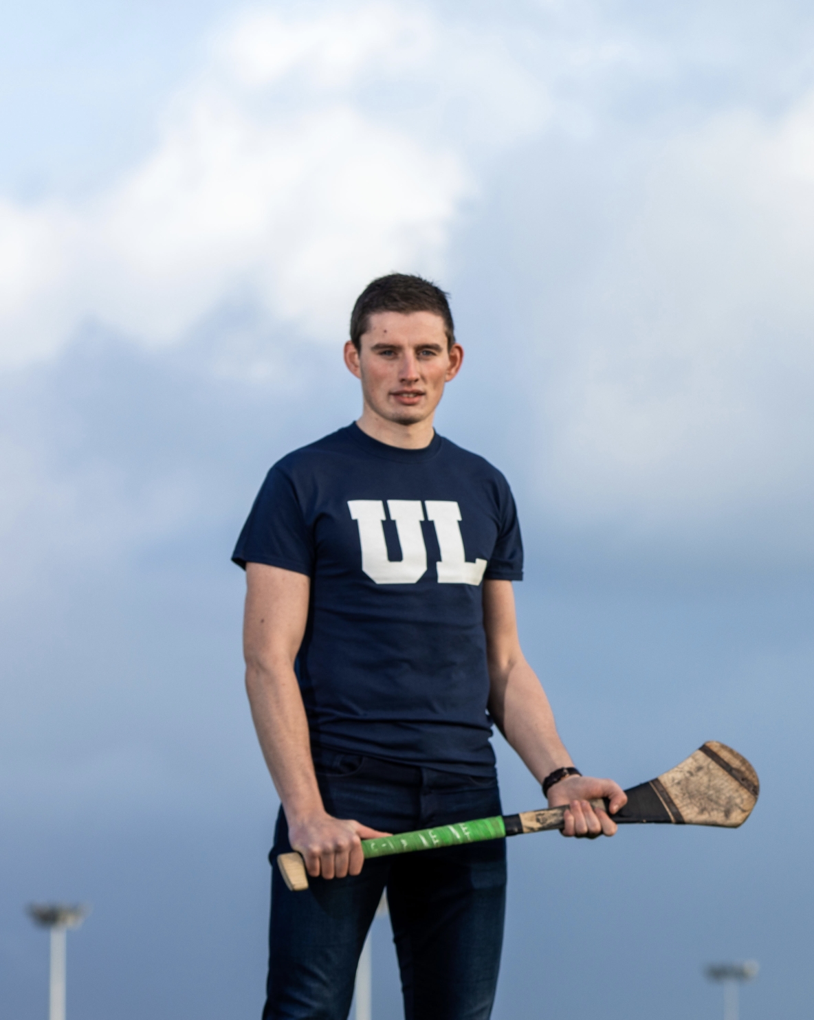 Gearoid Hegarty in a navy t-shirt with UL in white on the front.  He is holding a hurley in both hands and looking directly at the camera with clear sky behind him.
