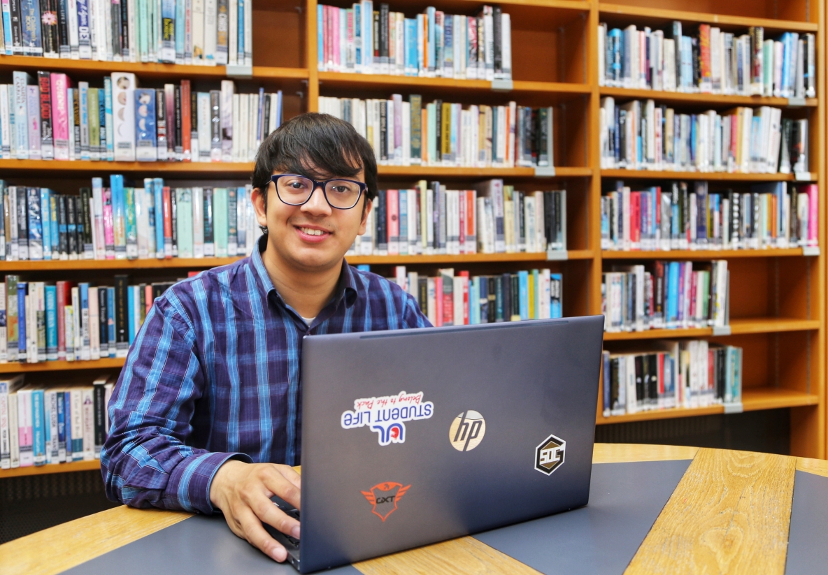 Picture of a student with a laptop at a desk, in the background is a bookshelf with a lot of books on it