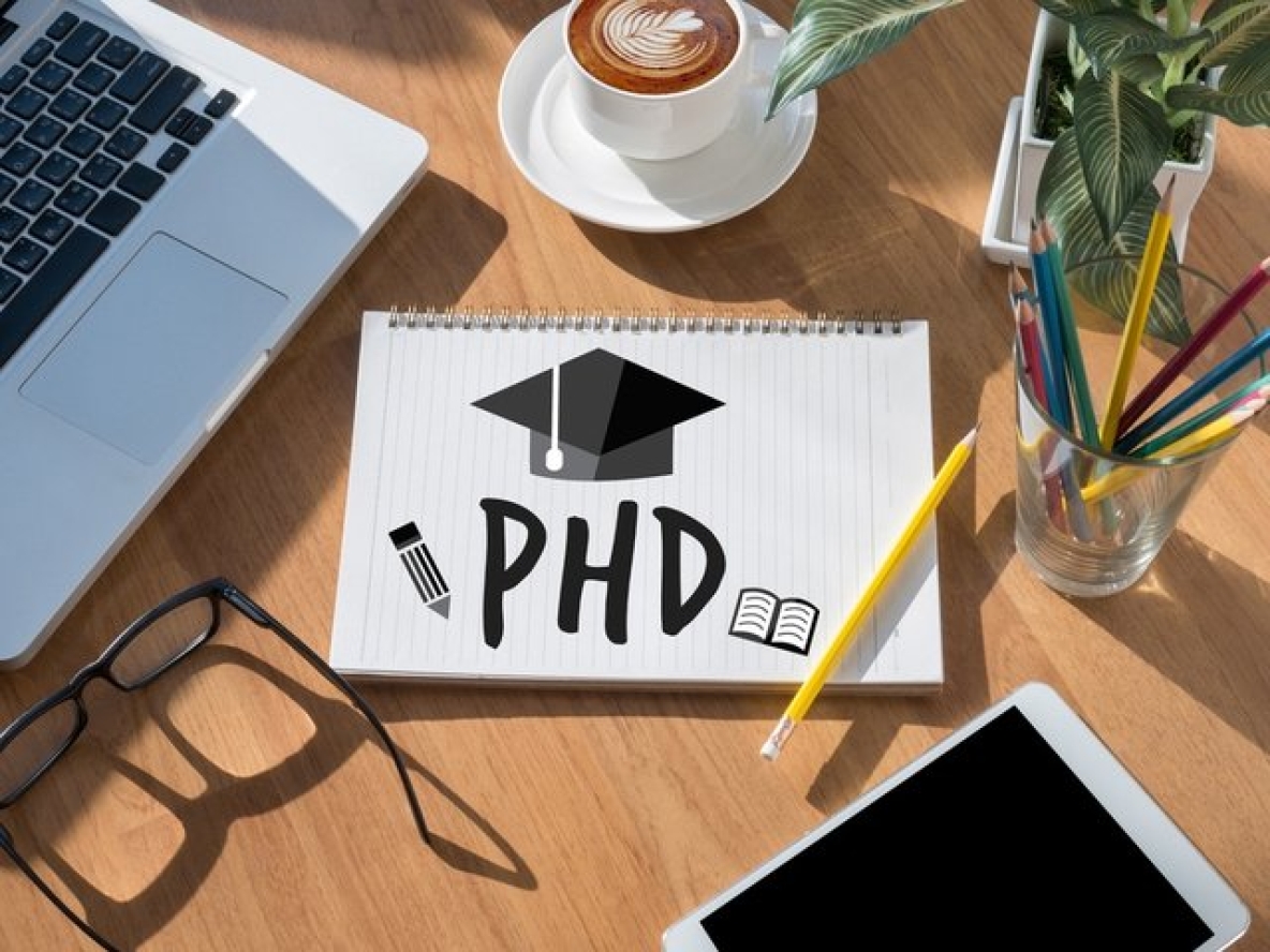 stock image of a laptop and notepad with PHD written on it and a graduation cap on a desk.
