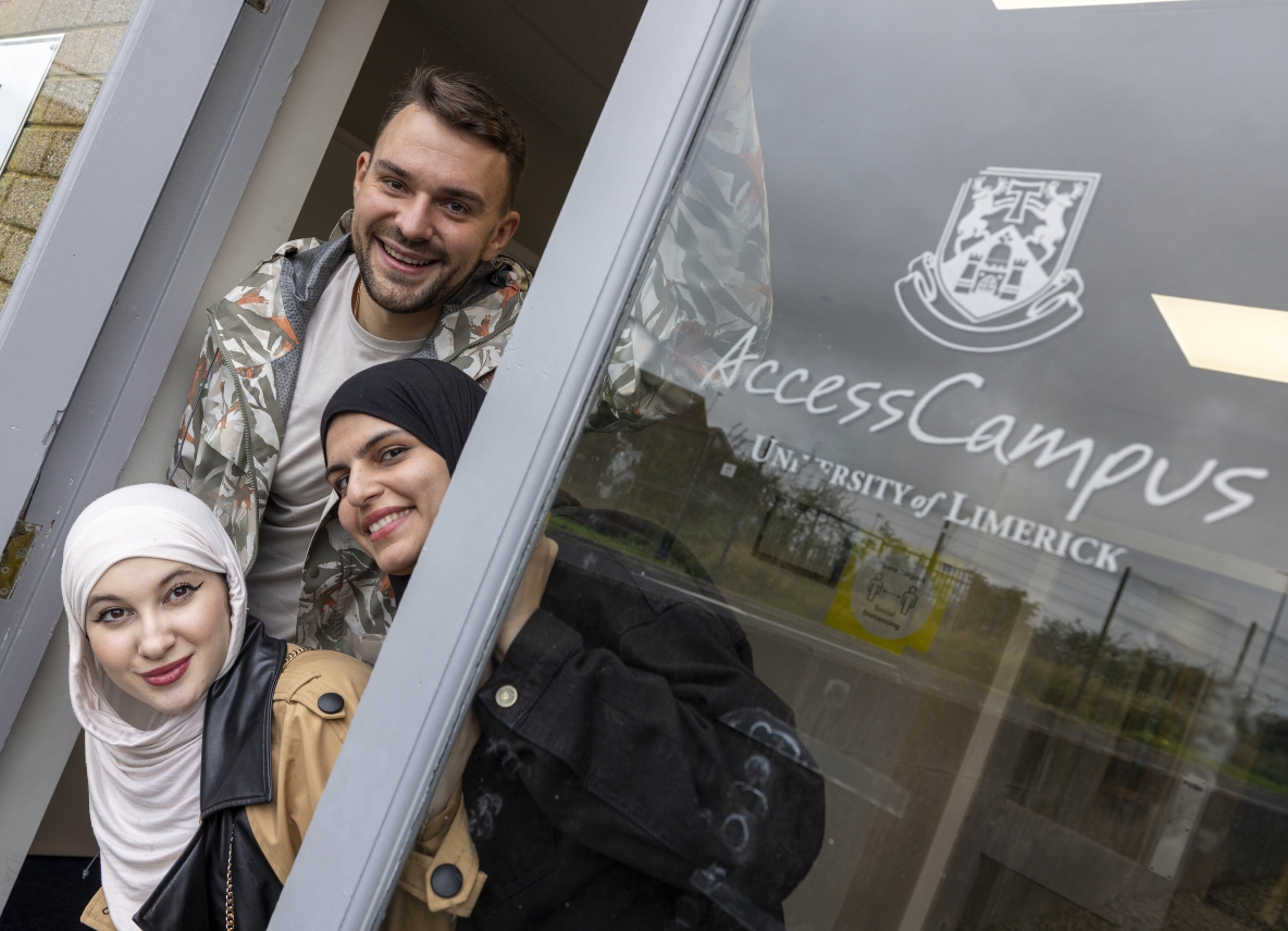 Two smiling female students wearing hijabs and a male student peaking out from the glass door with the sign UL Access Campus 