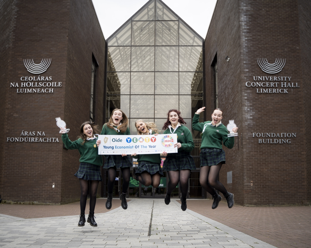 Five very happy teenage girls in green secondary school uniforms jumping for joy holding a poster which reads Young Economist of the Year