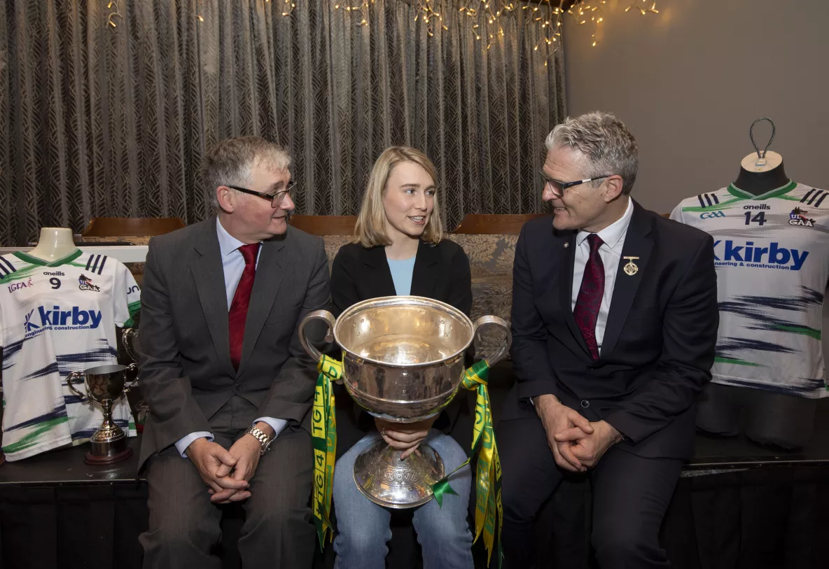 Three people sitting on a bench, two men and a woman. The woman is in the centre holding a large sports trophy..