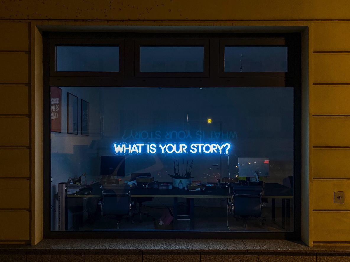 Image shows a window with the text "what is your story" in neon lights.