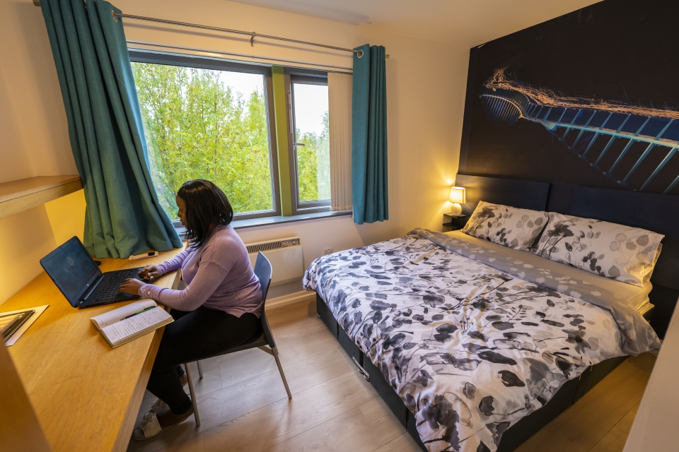 Female student sitting at desk, using laptop, in student accommodation