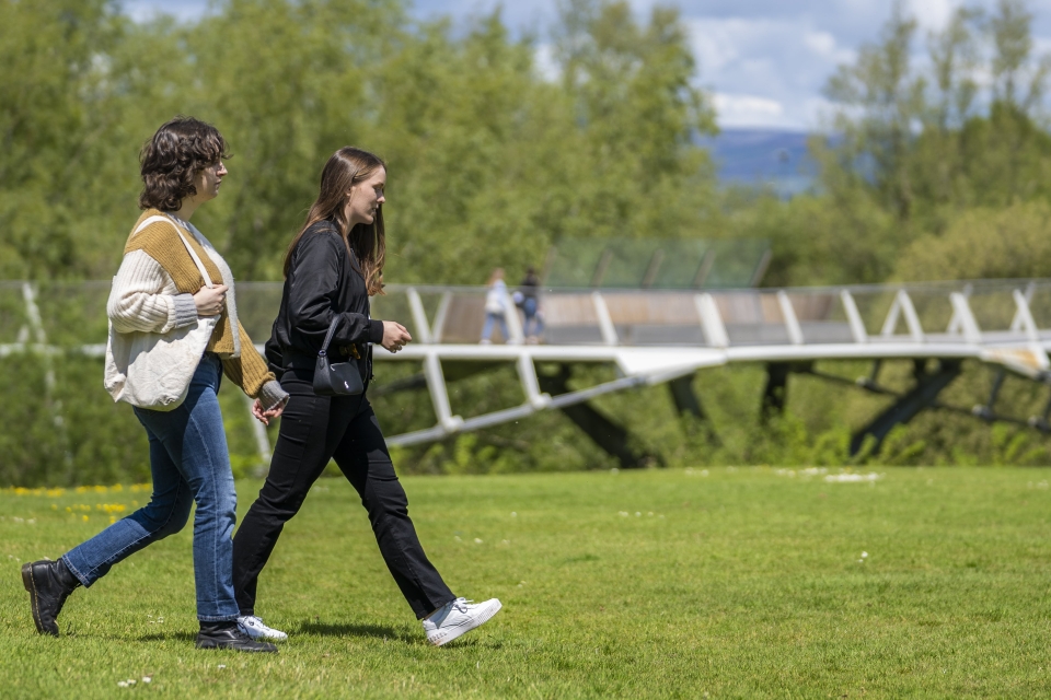 Two female students walking on a lawn with the Living Bridge in the background.