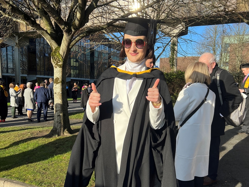 A woman in a black graduation cap and gown wearing sunglasses giving two thumbs up